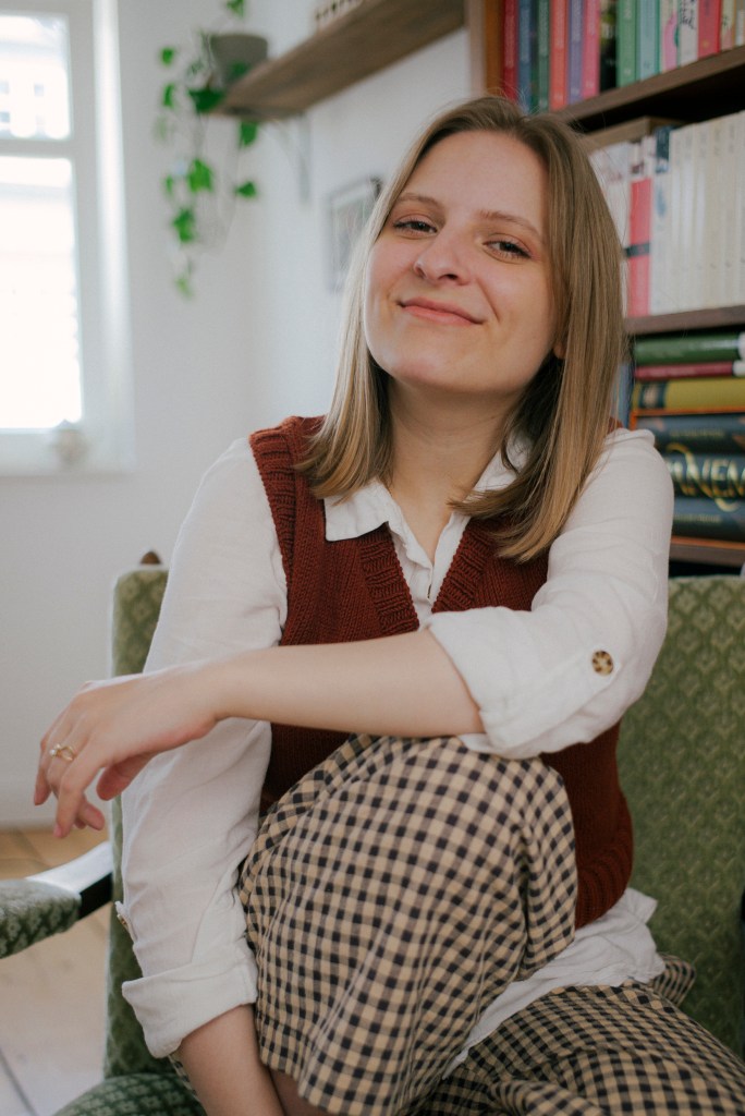 Auf diesem Foto ist Rebecca zu sehen. Sie lächelt verschmitzt und sitzt entspannt in einem grünen Sessel, der vor einem Bücherregal steht. 